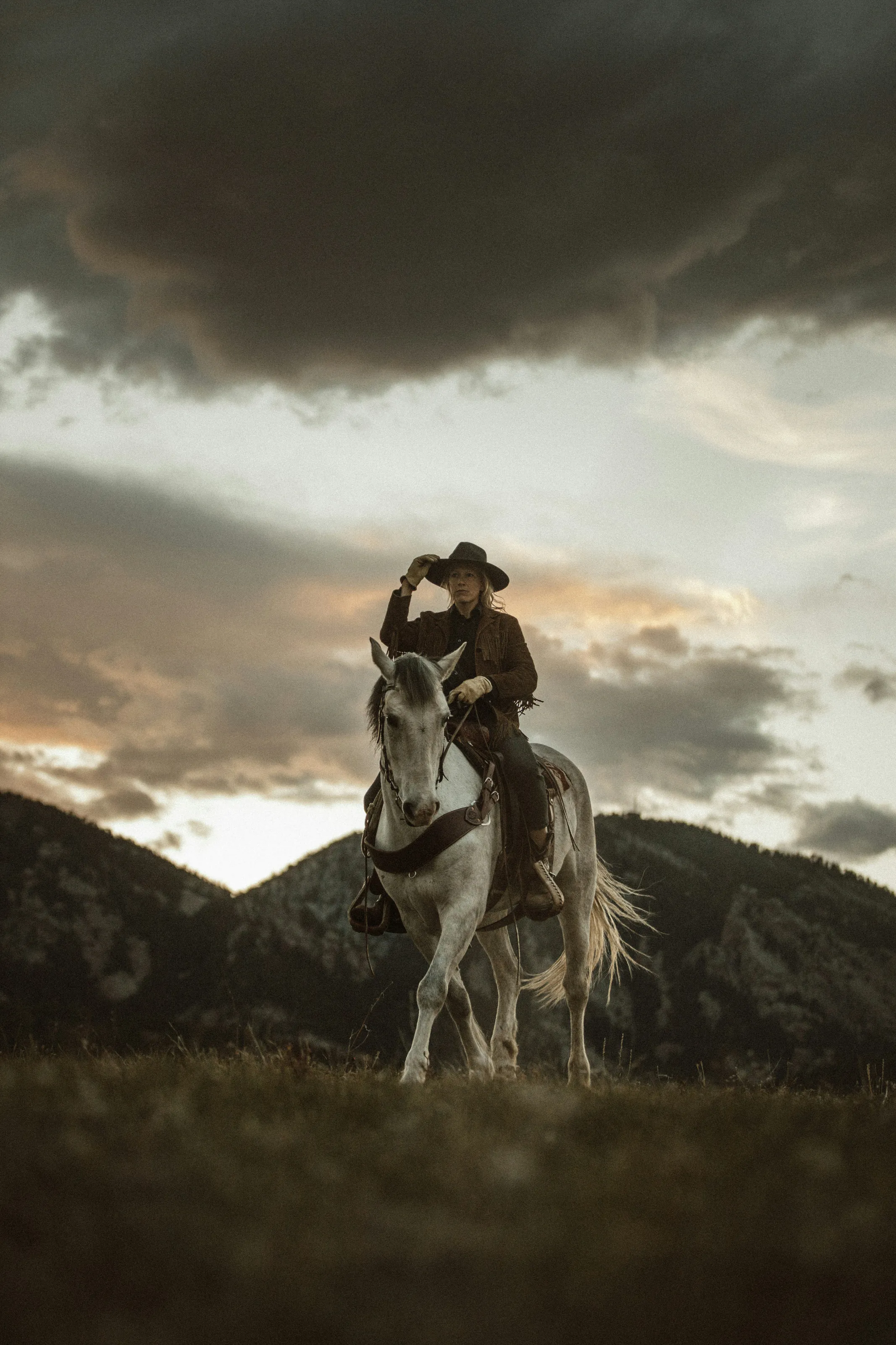 A rider in a wide-brimmed hat on a grey horse, hills and a storm sky behind